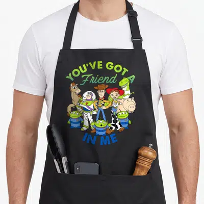 Cartoon dog breed aprons - Toy Story characters clustered below YOU'VE GOT A Friend IN ME text in green and blue — shown on a black cotton canvas apron against a white background