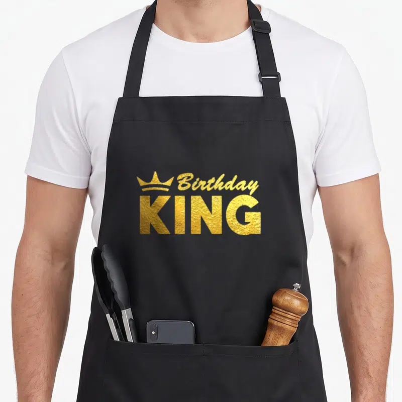 Crown squad kings apron - Gold crown icon beside Birthday KING text in textured metallic finish — shown on a black cotton canvas apron against a white background