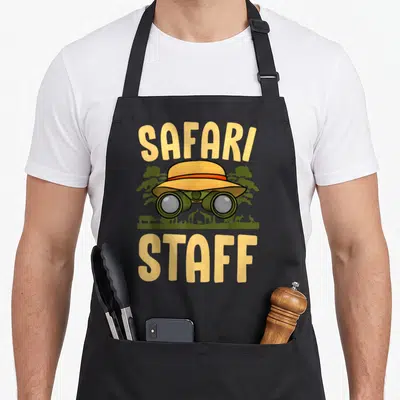 Safari staff aprons - SAFARI STAFF text with pith hat, binoculars, and animal silhouettes on green ground line — shown on a black cotton canvas apron against a white background