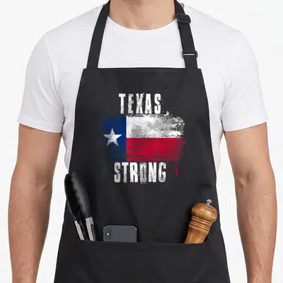 Strong texans apron - Distressed Texas flag with white TEXAS STRONG lettering in blue, white, and red — shown on a black cotton canvas apron against a white background