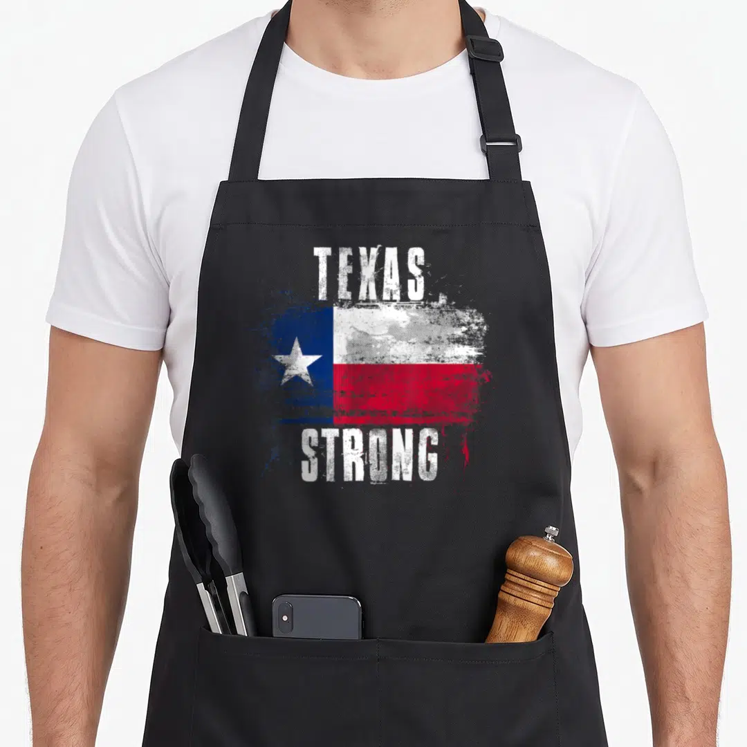 Strong texans apron - Distressed Texas flag with white TEXAS STRONG lettering in blue, white, and red — shown on a black cotton canvas apron against a white background