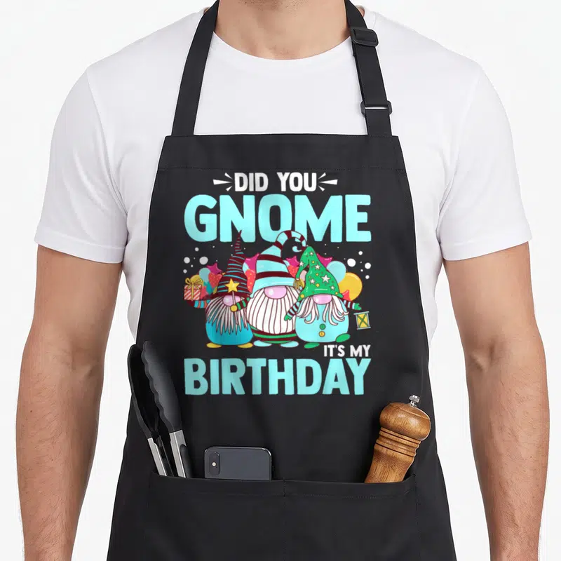 Gnome birthday apron - Three cartoon gnomes with balloons and gift under Did You Gnome It's My Birthday text — shown on a black cotton canvas apron against a white background