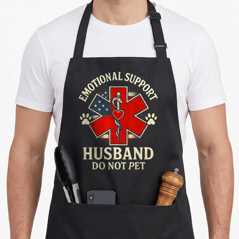 Flag husband apron - Red Star of Life with Rod of Asclepius and US flag behind EMOTIONAL SUPPORT HUSBAND text — shown on a black cotton canvas apron against a white background