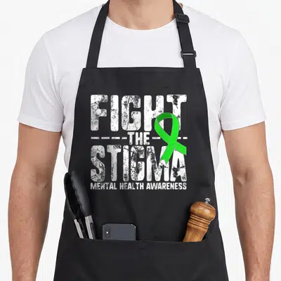 Fight spread apron - Distressed white text reading Fight The Stigma Mental Health Awareness with overlapping green — shown on a black cotton canvas apron against a white background