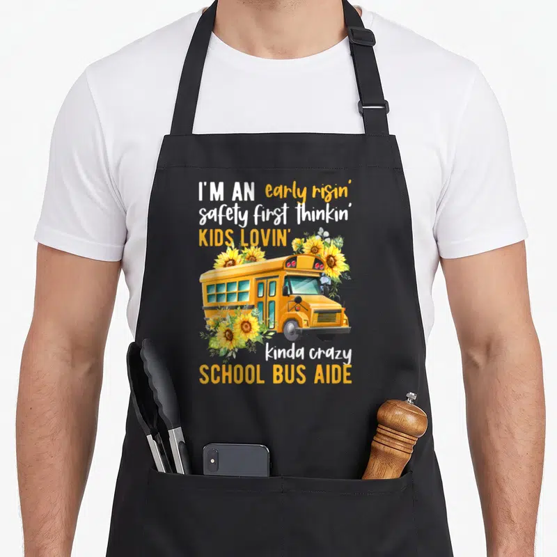 Aide school bus apron - Cartoon yellow bus with sunflowers and text reading I'm an early risin school bus aide — shown on a black cotton canvas apron against a white background