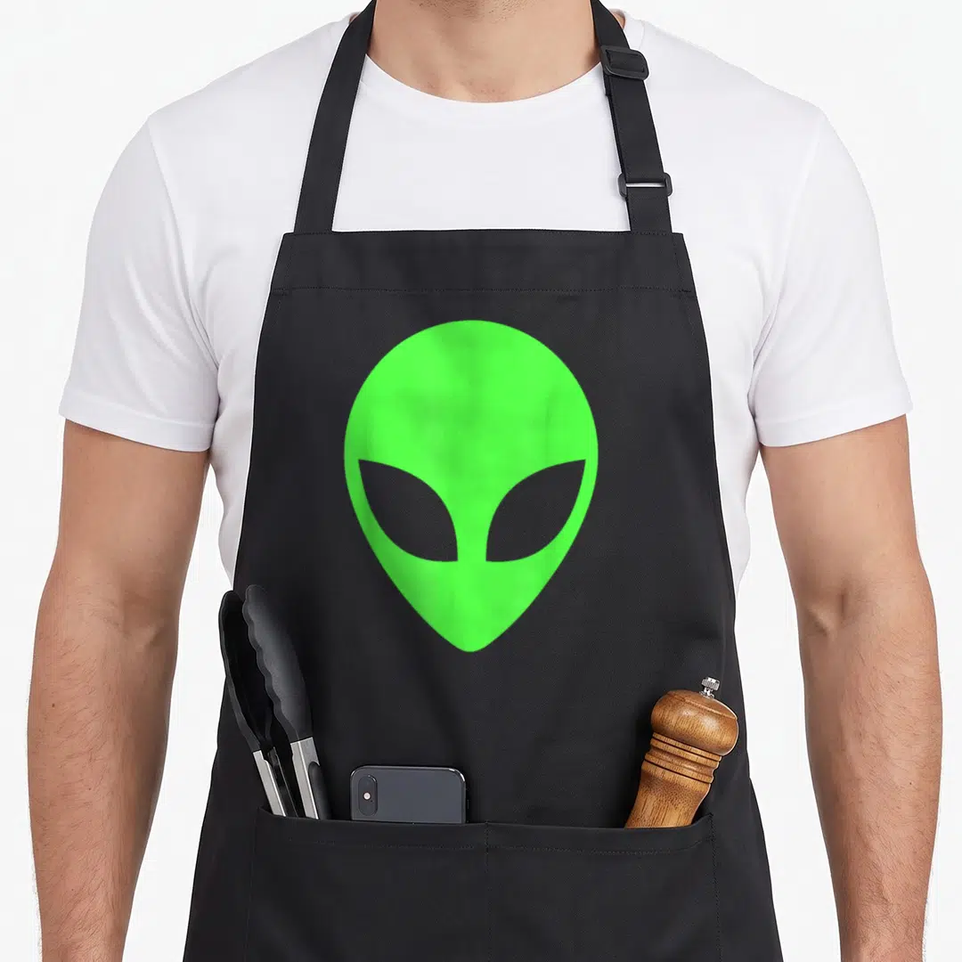 Cute funny alien apron - Lime green minimalist alien head graphic centered on fabric with two front pockets — shown on a black cotton canvas apron against a white background