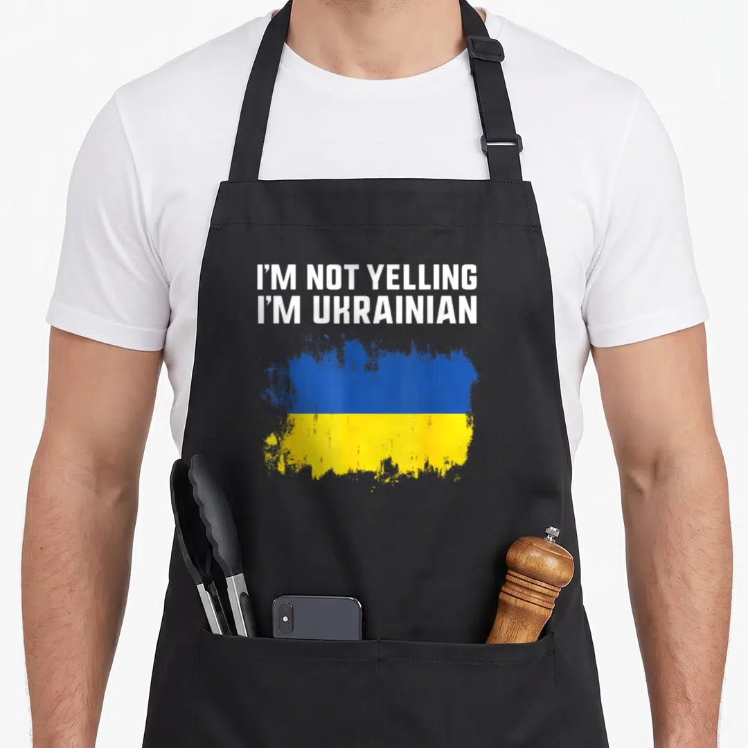 Pride yelling ukrainian aprons - White text I'm Not Yelling I'm Ukrainian above distressed blue and yellow flag — shown on a black cotton canvas apron against a white background