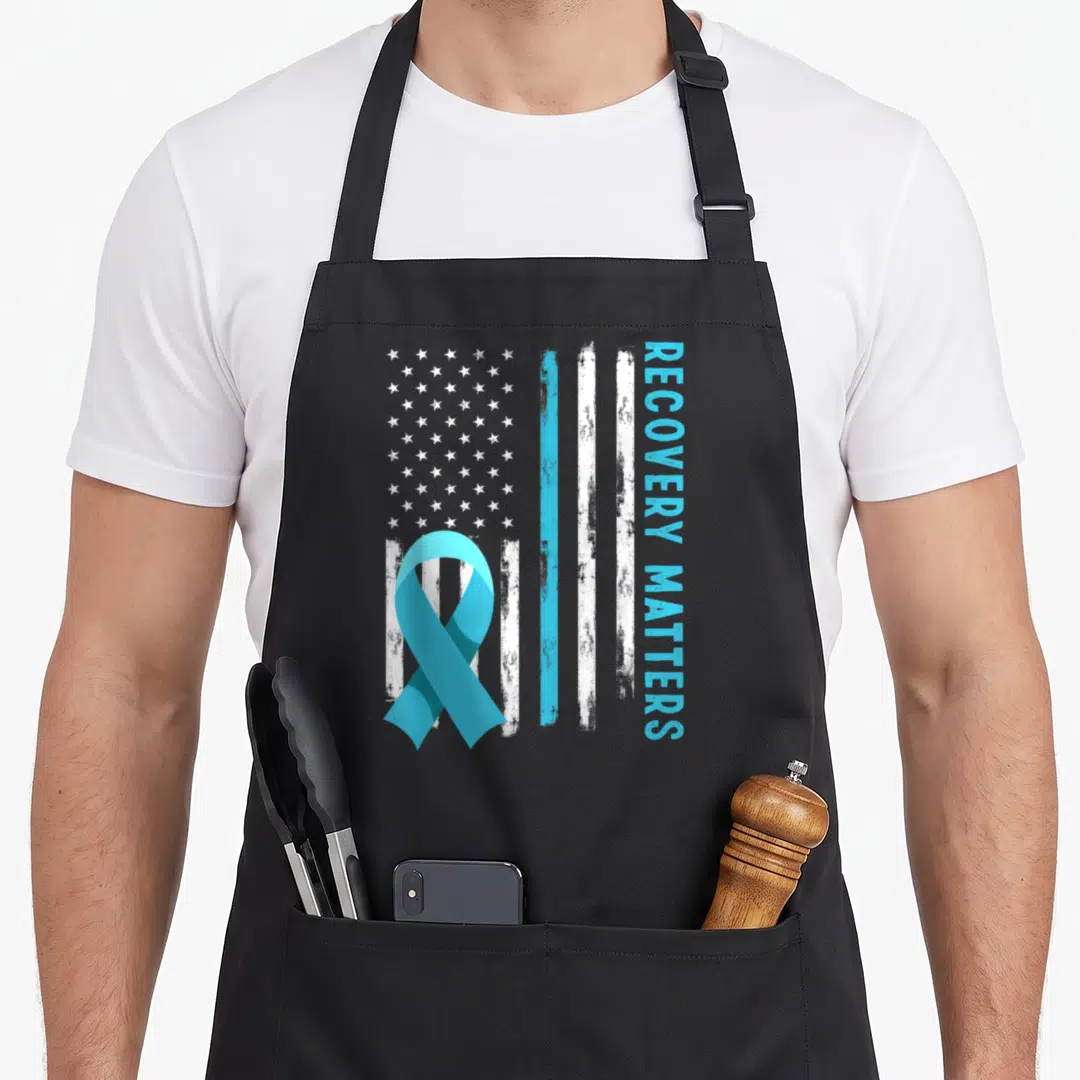 Alcohol american flag apron - Distressed flag with light blue ribbon and vertical RECOVERY MATTERS text — shown on a black cotton canvas apron against a white background