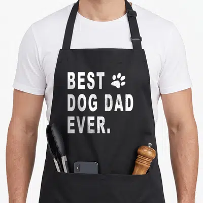Cute dog dad apron - White distressed text BEST DOG DAD EVER with paw print graphic — shown on a black cotton canvas apron against a white background