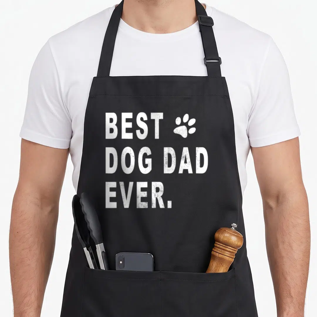 Cute dog dad apron - White distressed text BEST DOG DAD EVER with paw print graphic — shown on a black cotton canvas apron against a white background