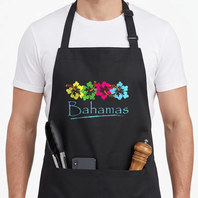 Bahamas tropical print aprons - Four yellow green pink and blue hibiscus flowers above light blue Bahamas text — shown on a black cotton canvas apron against a white background