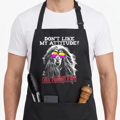 Afghan attitude aprons - Long-haired dog with yellow and pink sunglasses and sarcastic text about dogs versus cats — shown on a black cotton canvas apron against a white background