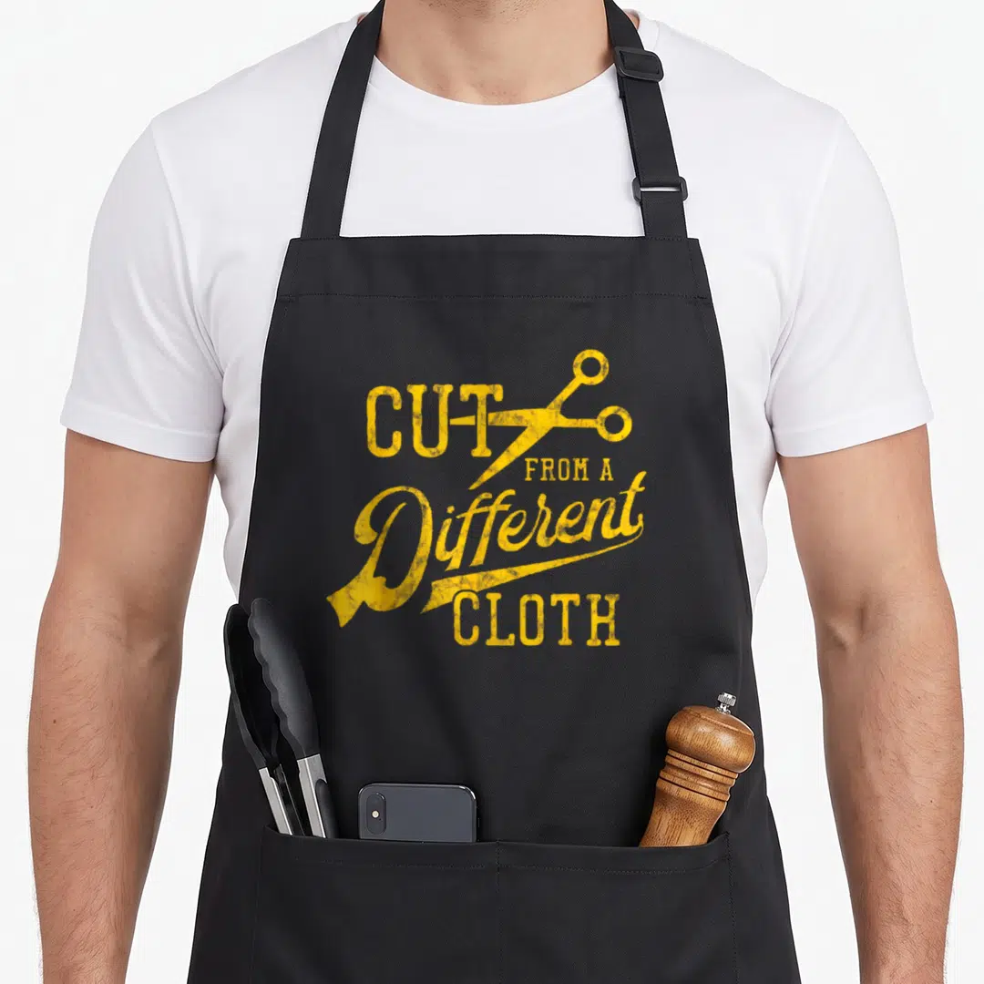 Cut hip aprons - Distressed yellow text CUT FROM A Different CLOTH with scissors icon — shown on a black cotton canvas apron against a white background
