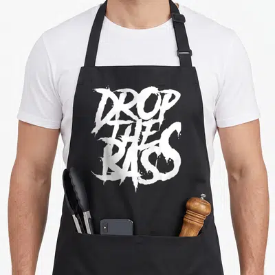 Drop dubstep apron - White stacked text reading DROP THE BASS in stylized brush font on grey background — shown on a black cotton canvas apron against a white background