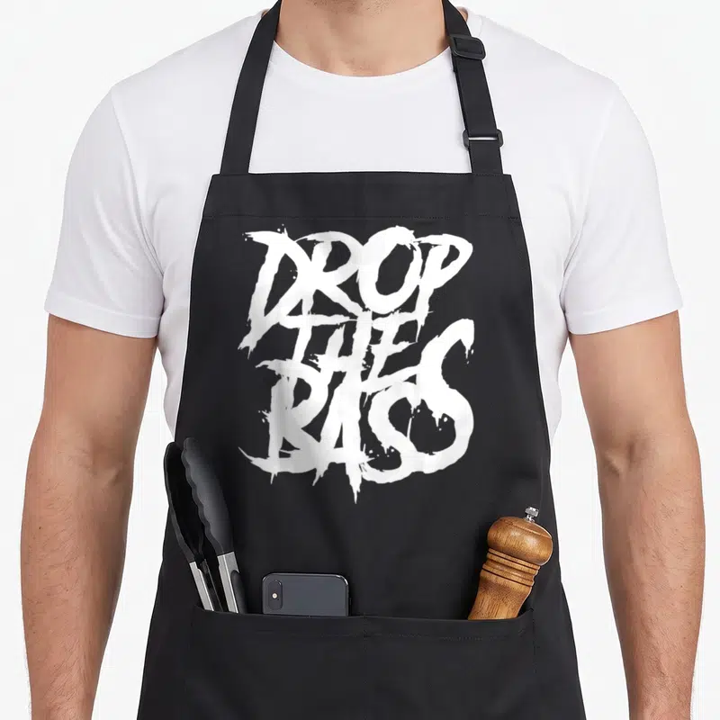 Drop dubstep apron - White stacked text reading DROP THE BASS in stylized brush font on grey background — shown on a black cotton canvas apron against a white background