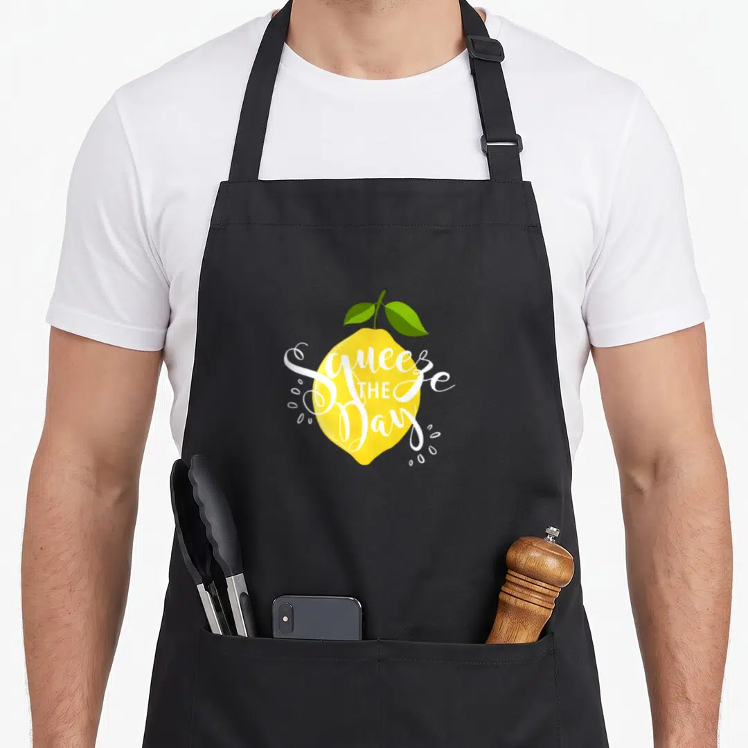 Squeeze lemon apron - Yellow lemon with Squeeze THE Day text, green leaves, and white juice drops — shown on a black cotton canvas apron against a white background