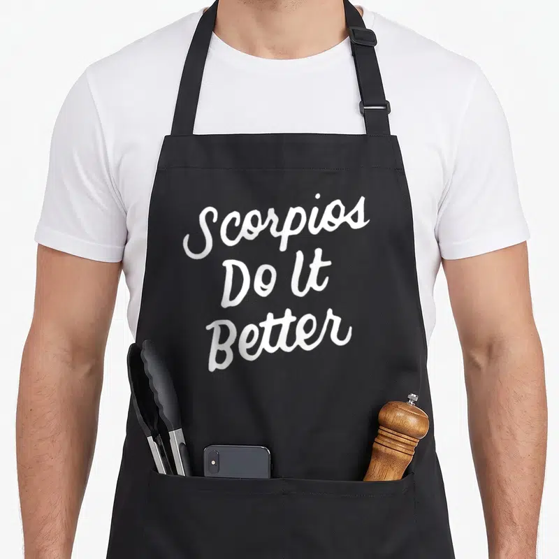 Better zodiac aprons - White stacked script text reading Scorpios Do It Better on black fabric — shown on a black cotton canvas apron against a white background