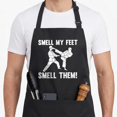 Funny karate martial apron - White line art of two martial artists kicking with Smell My Feet text — shown on a black cotton canvas apron against a white background