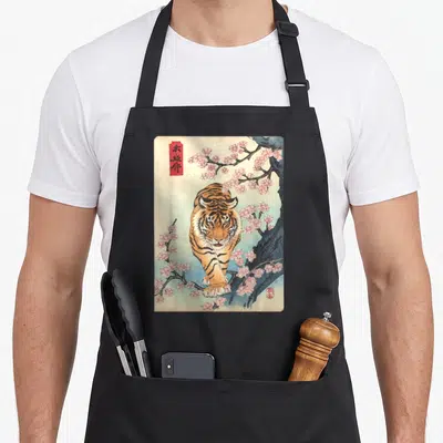 Asian cherry apron - Vintage tiger walking under pink cherry blossoms with red artist seals and blue wash — shown on a black cotton canvas apron against a white background