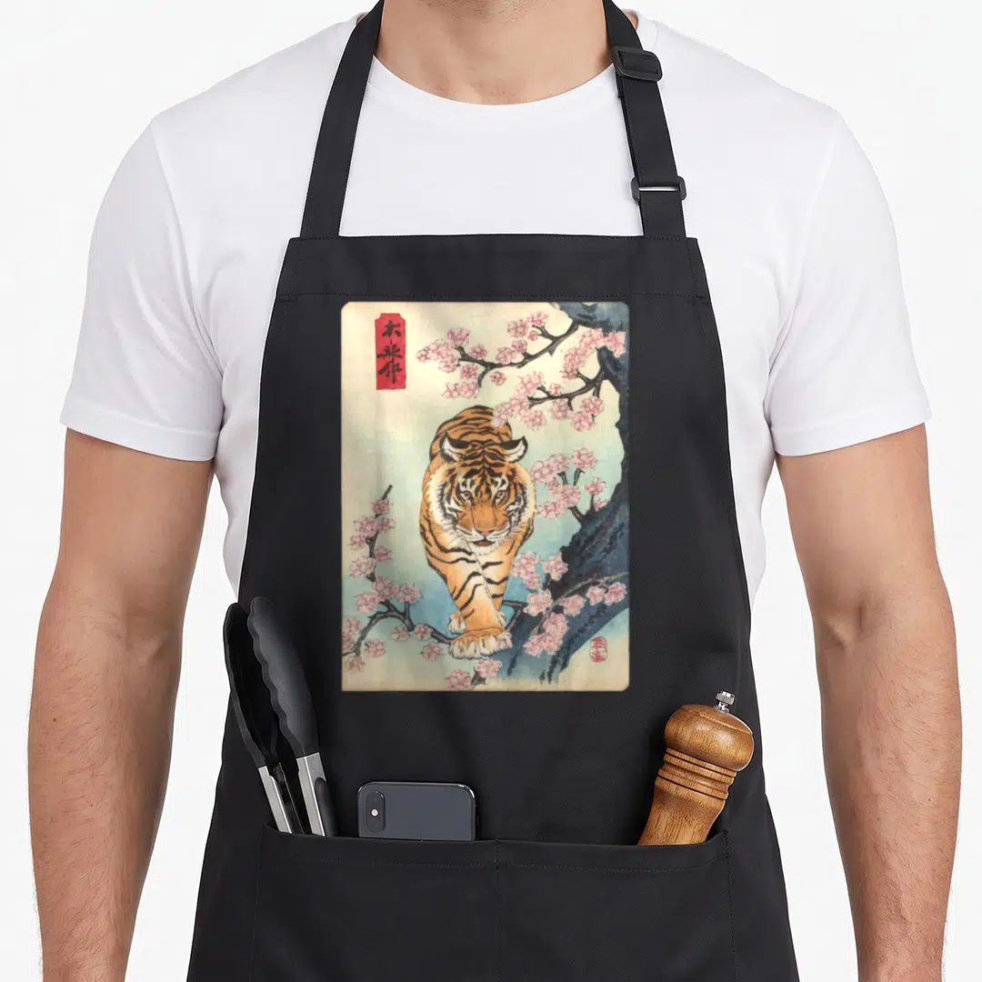 Asian cherry apron - Vintage tiger walking under pink cherry blossoms with red artist seals and blue wash — shown on a black cotton canvas apron against a white background