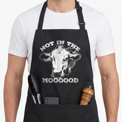 Mood cow apron - Distressed white cow head with arched text NOT IN THE and straight text MOOOOOD below — shown on a black cotton canvas apron against a white background