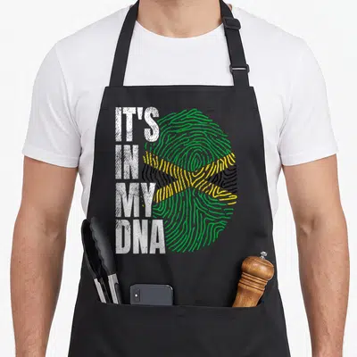 Jamaican pride apron - Fingerprint in green yellow black and white with text IT'S IN MY DNA — shown on a black cotton canvas apron against a white background