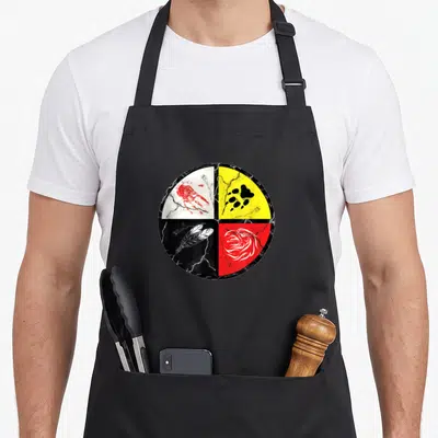 Medicine wheel apron - Distressed circle with four colored quadrants showing handprint, paw, feather, and eagle head — shown on a black cotton canvas apron against a white background