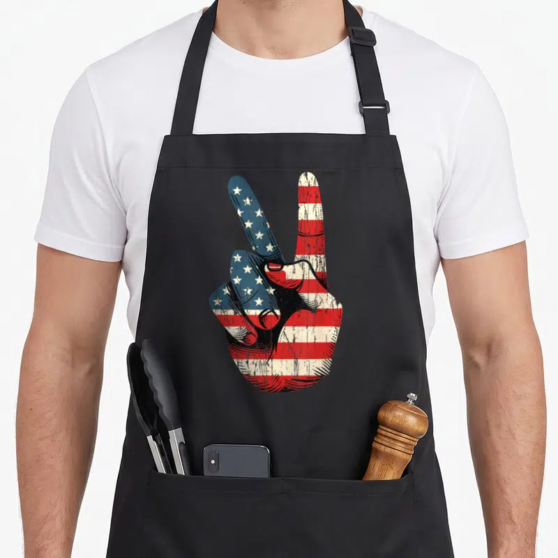 Peace sign 60s apron - Distressed hand making peace sign filled with USA flag pattern in red white and blue — shown on a black cotton canvas apron against a white background