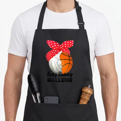 Baller busy apron - Split volleyball basketball with red polka dot bow and Busy Raising Ballers text — shown on a black cotton canvas apron against a white background