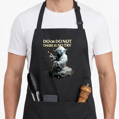 Try apron or apron - Yoda sitting on rock with Do Or Do Not There Is No Try text and gold sparkles — shown on a black cotton canvas apron against a white background
