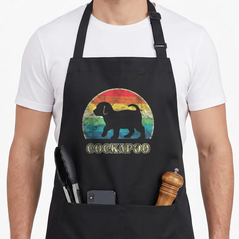Cool dog cockapoo apron - Grey dog silhouette over red orange yellow green blue striped arch with COCKAPOO text — shown on a black cotton canvas apron against a white background