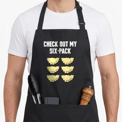 Pierogi polish aprons - Six yellow dumplings in two columns under white CHECK OUT MY SIX-PACK text — shown on a black cotton canvas apron against a white background