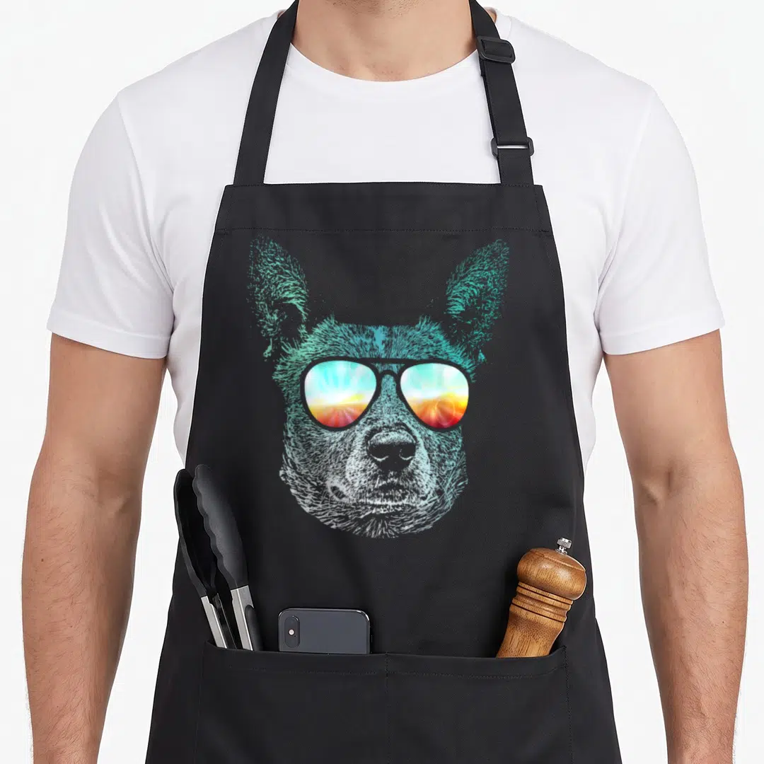 Stumpy dog kitchen apron - Realistic blue dog head wearing sunglasses reflecting a sunset in light blue, white, red — shown on a black cotton canvas apron against a white background