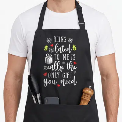 Related family christmas aprons - White stacked text reading BEING related TO ME IS really the ONLY GIFT you need — shown on a black cotton canvas apron against a white background