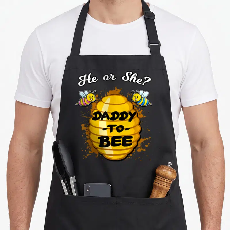 Daddy bee apron - Cartoon beehive with He or She text and two flanking bees in orange splashes — shown on a black cotton canvas apron against a white background