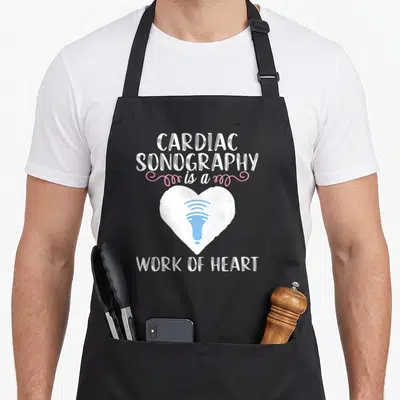 Ultrasound sonographer apron - White heart with blue transducer and pink swirls surrounding stacked text — shown on a black cotton canvas apron against a white background