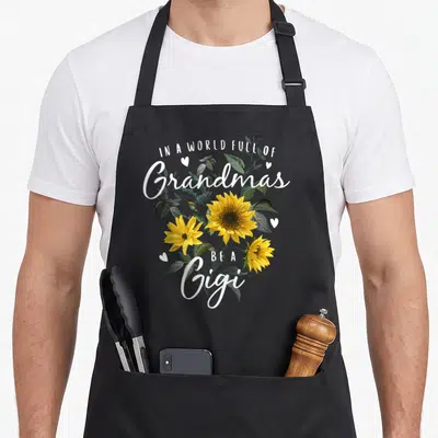Sunflower grandma's aprons - Yellow sunflowers and white text reading Be A Gigi surrounded by small hearts — shown on a black cotton canvas apron against a white background