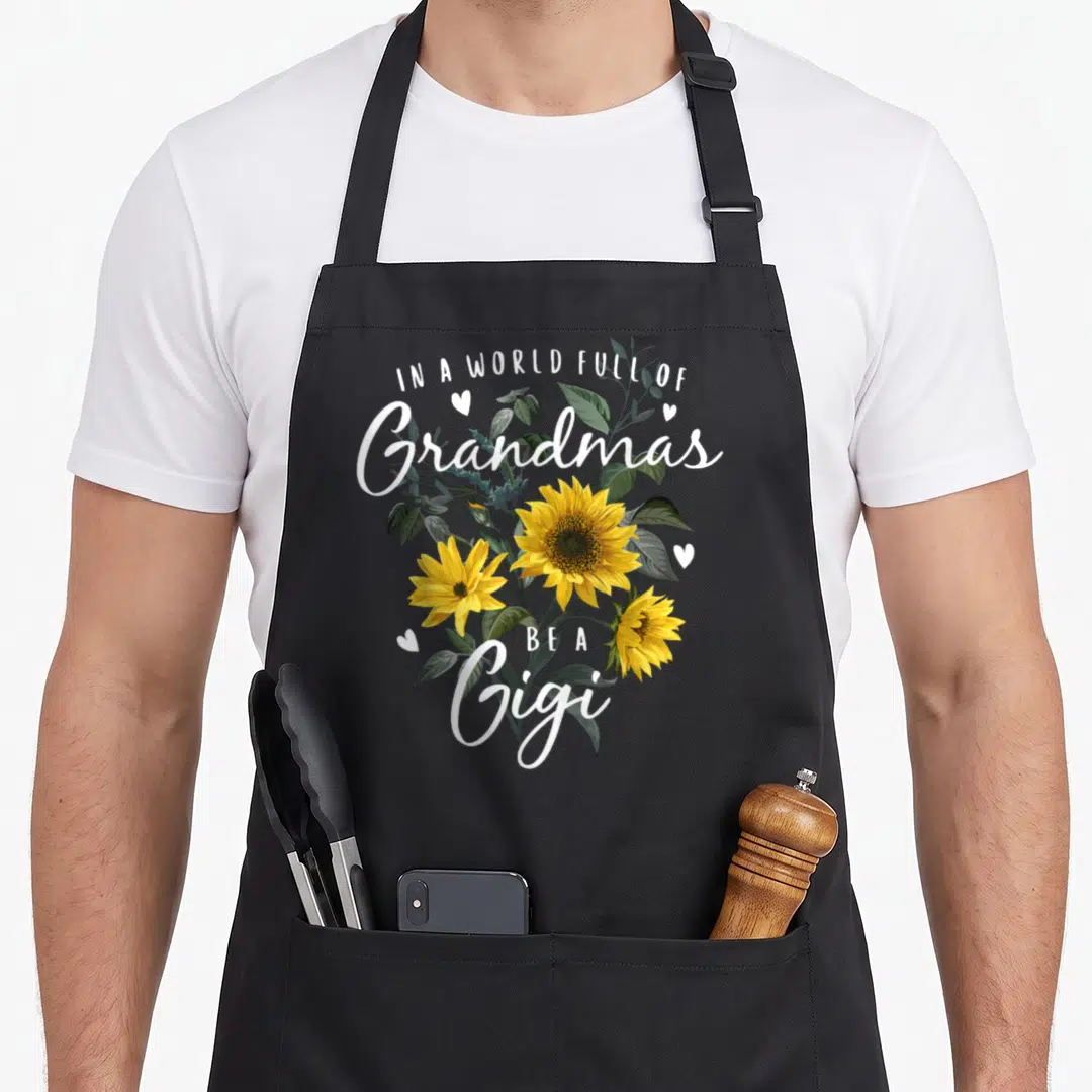 Sunflower grandma's aprons - Yellow sunflowers and white text reading Be A Gigi surrounded by small hearts — shown on a black cotton canvas apron against a white background