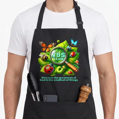 Magnified christian apron - Cartoon magnifying glass over leaves and insects with VBS TEAM text and inspirational quote — shown on a black cotton canvas apron against a white background
