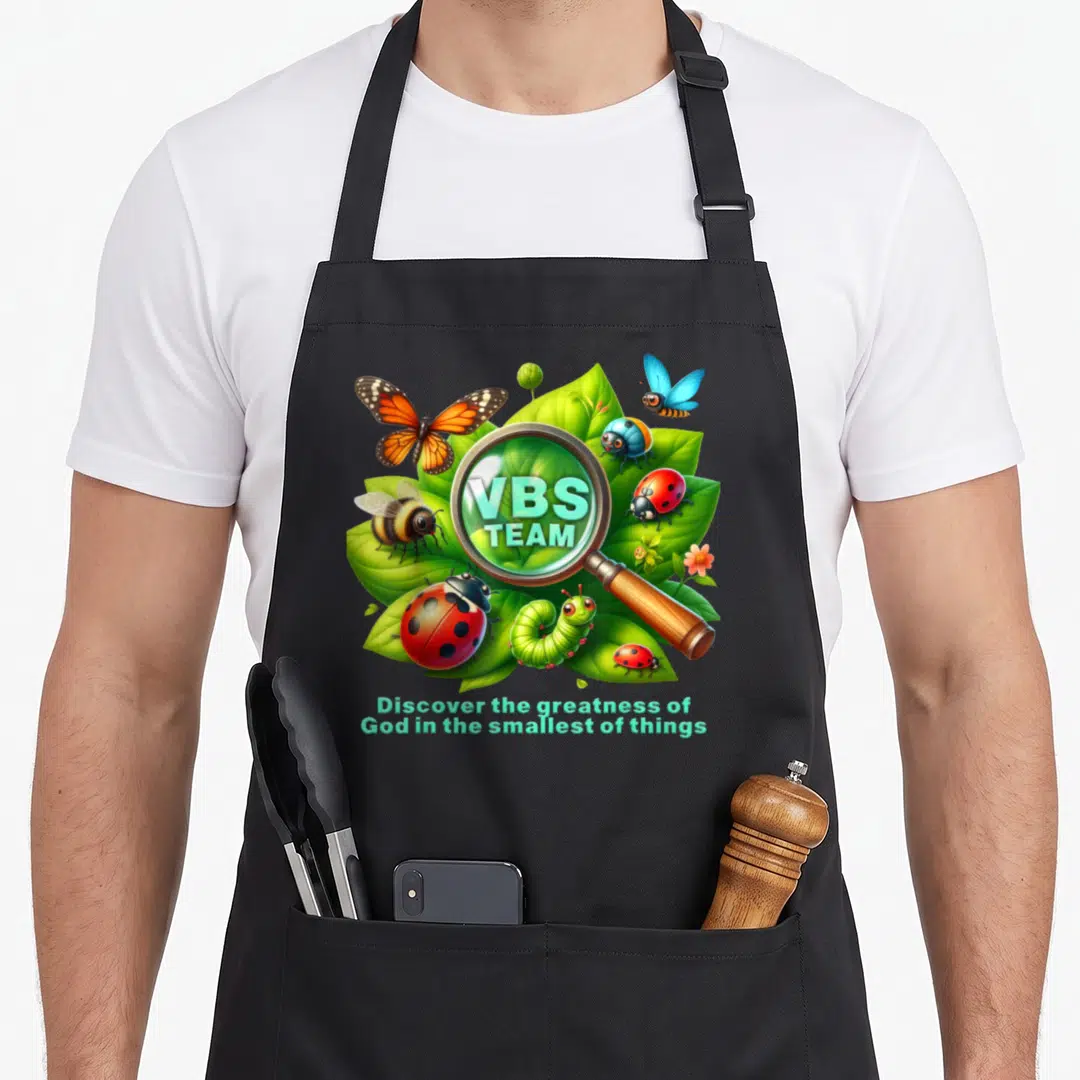 Magnified christian apron - Cartoon magnifying glass over leaves and insects with VBS TEAM text and inspirational quote — shown on a black cotton canvas apron against a white background
