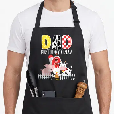 Crew farm apron - DAD BIRTHDAY CREW text above red barn, cow, pig, chicken, and white picket fence illustration — shown on a black cotton canvas apron against a white background