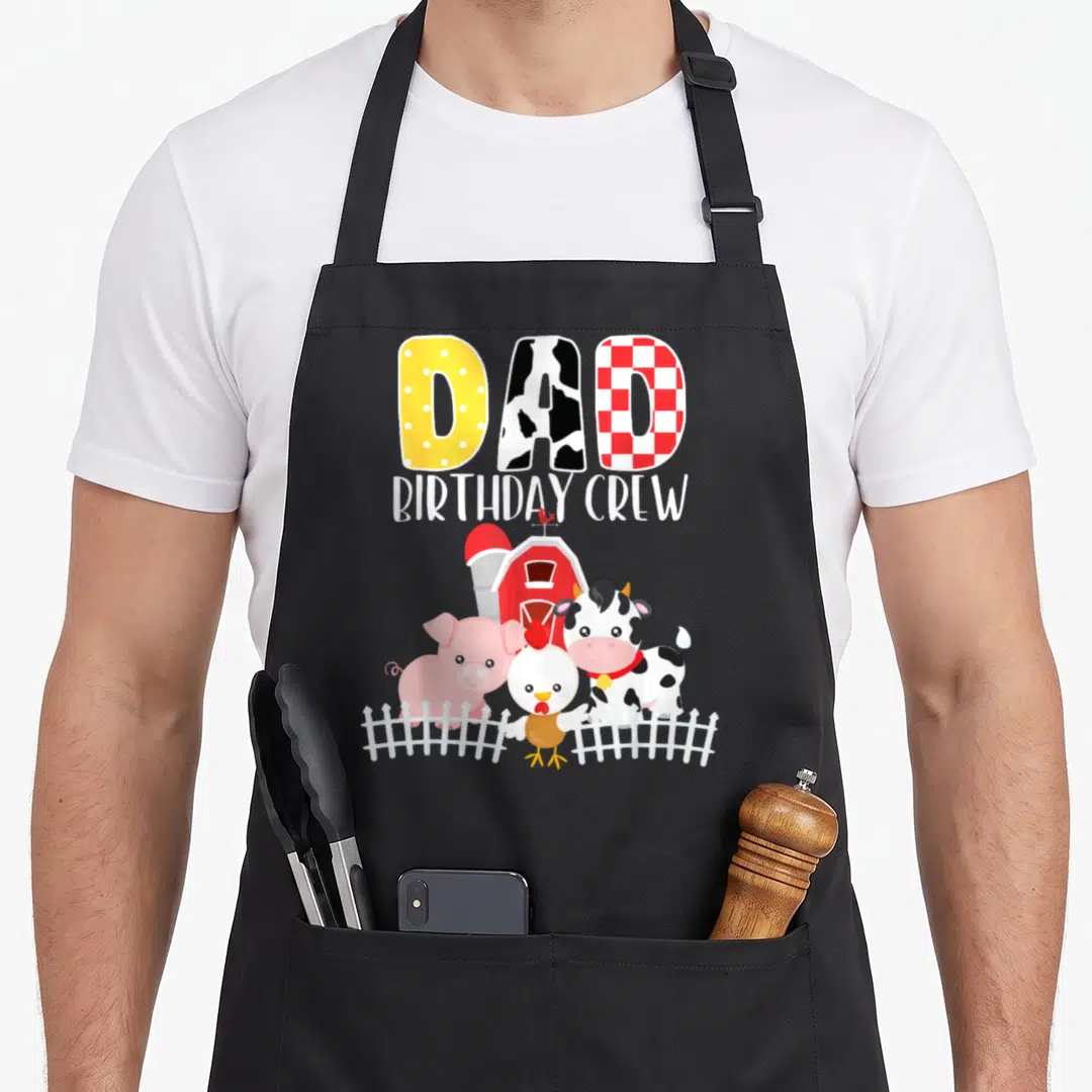 Crew farm apron - DAD BIRTHDAY CREW text above red barn, cow, pig, chicken, and white picket fence illustration — shown on a black cotton canvas apron against a white background