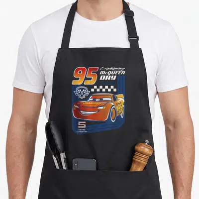 Trading car apron - Red cartoon race car with 95, Lightning McQueen Day text, checkered flags, and 5 TIME CHAMP on blue — shown on a black cotton canvas apron against a white background