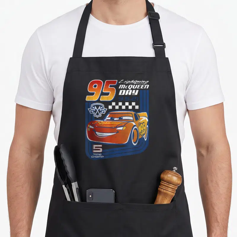 Trading car apron - Red cartoon race car with 95, Lightning McQueen Day text, checkered flags, and 5 TIME CHAMP on blue — shown on a black cotton canvas apron against a white background