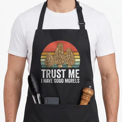 Mushroom hunter foraging apron - Three brown morels over striped sun with white text TRUST ME I HAVE GOOD MORELS — shown on a black cotton canvas apron against a white background