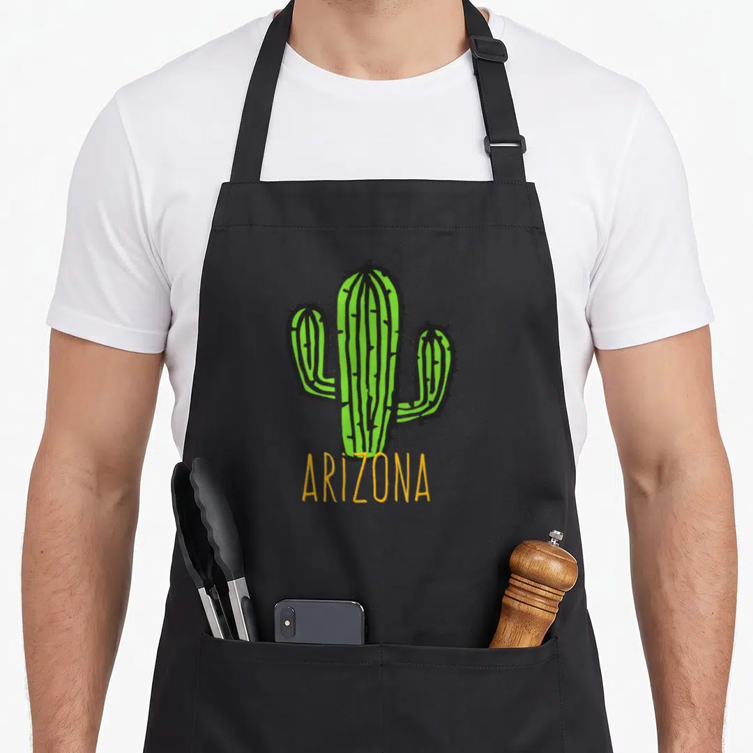Arizona souvenir cactus apron - Green cartoon saguaro with yellow ARIZONA text below — shown on a black cotton canvas apron against a white background