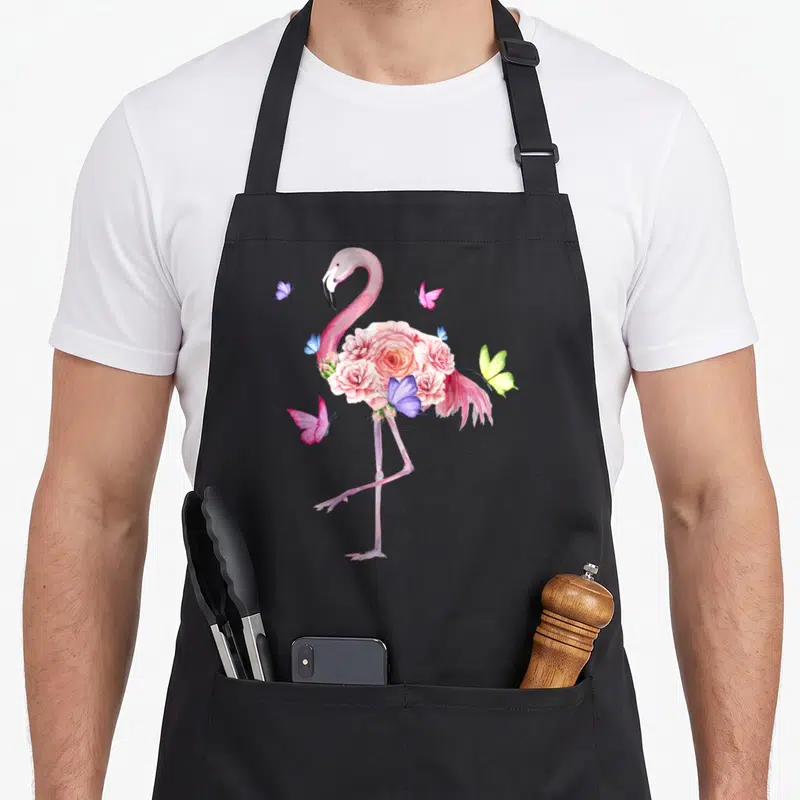 Nature pink flamingo apron - Watercolor pink flamingo with rose cluster midsection and scattered blue, yellow — shown on a black cotton canvas apron against a white background
