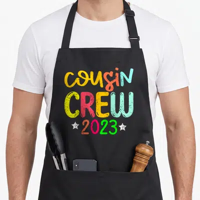 Cousin crew memory apron - Stacked text reading cousin CREW 2023 in yellow orange red blue green pink with two white — shown on a black cotton canvas apron against a white background