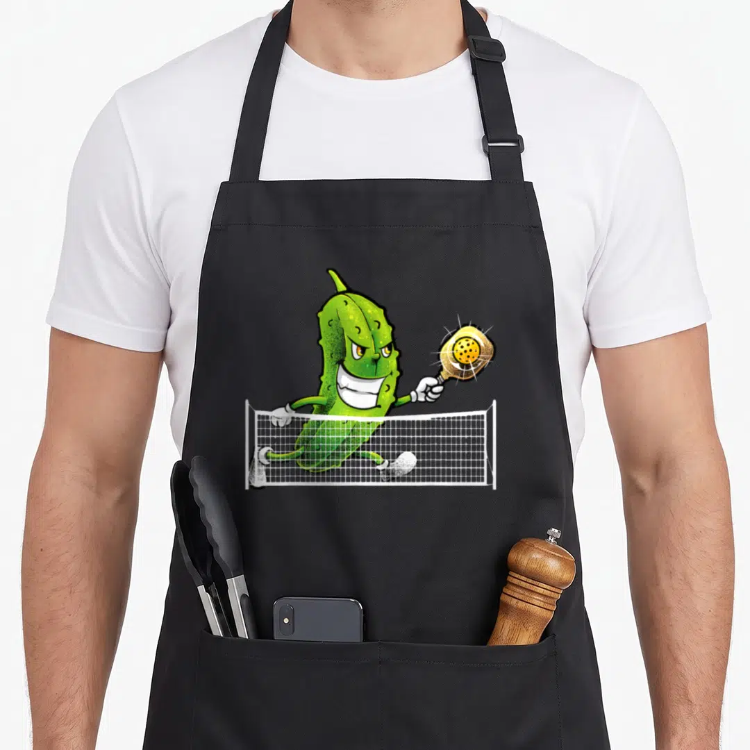 Racket pickleball apron - Cartoon green pickle running behind white net holding yellow paddle — shown on a black cotton canvas apron against a white background