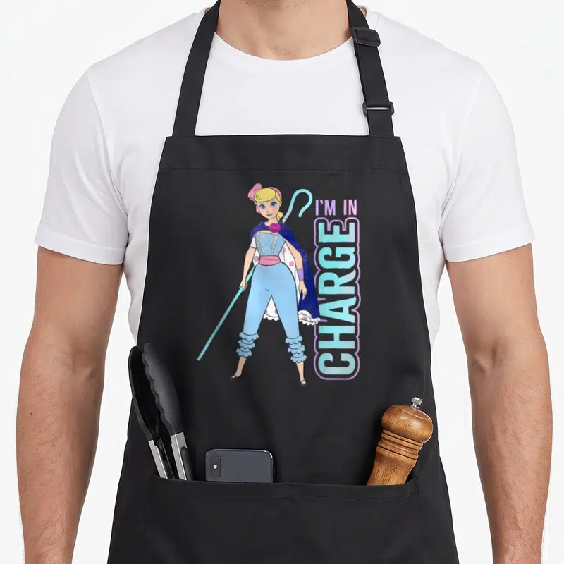 Charge bo peep apron - Cartoon woman with crook and vertical I'M IN CHARGE text in blue purple pink — shown on a black cotton canvas apron against a white background